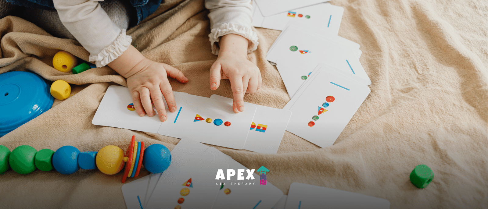 Toddler's hands sorting cards with colorful shapes, surrounded by wooden beads and toys on a soft blanket.