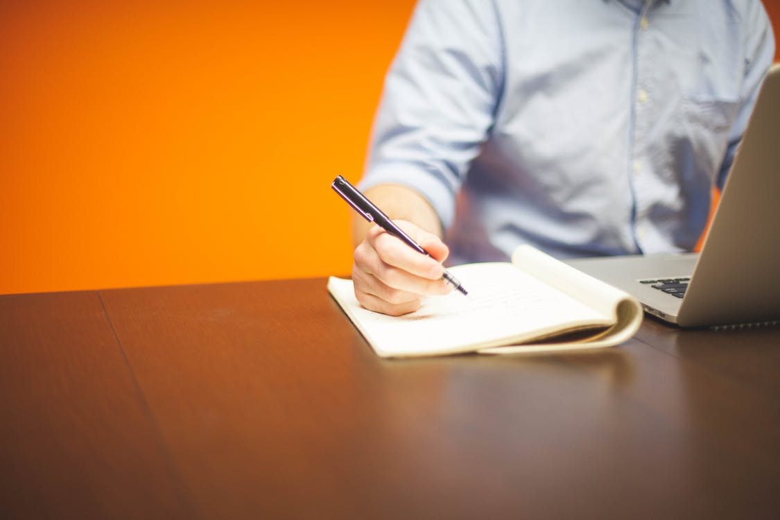 Free Person Writing on Note Beside Gray Laptop Computer Stock Photo
