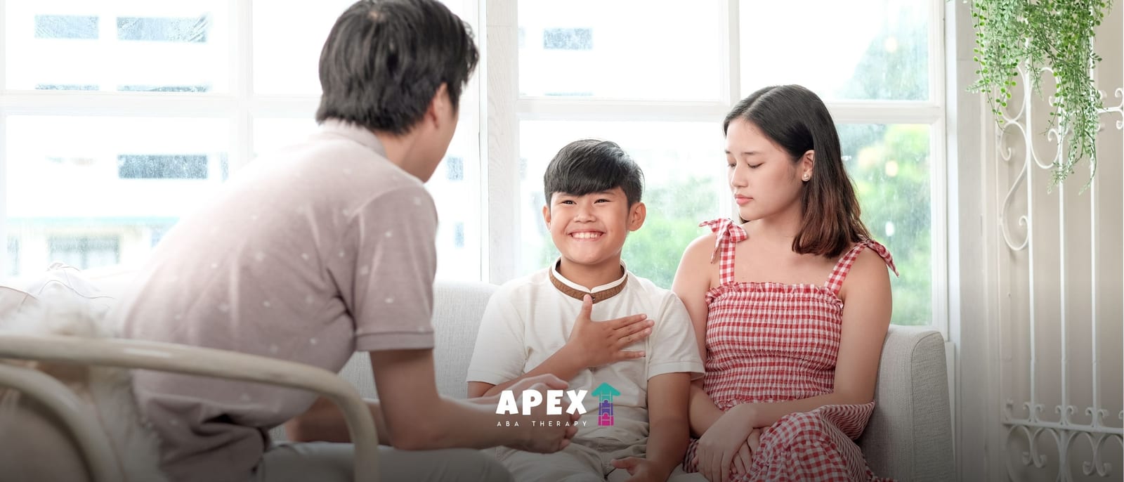 In a bright living room, a smiling boy sits between a man and woman, hand on his chest as they talk in a calm, supportive family conversation.