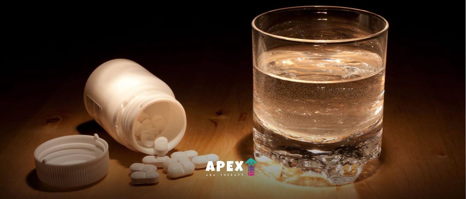 An overturned white pill bottle spills tablets beside a glass of water on a wooden surface, dramatically lit against a dark background; the mood is somber and cautionary.