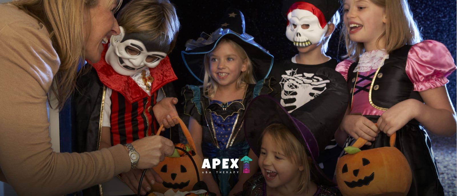 An adult helps costumed children gather around carved pumpkins: a witch, vampire, skeleton and princess smile in a cheerful Halloween scene.