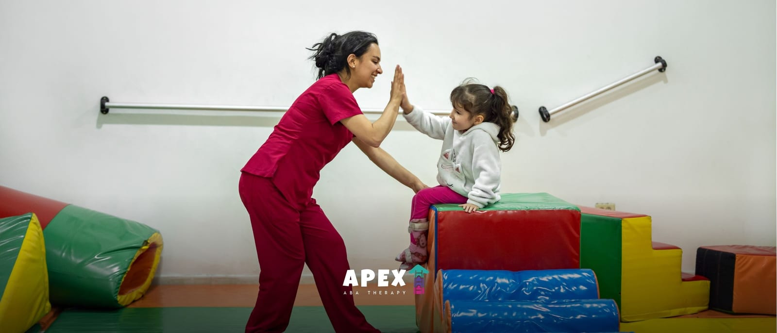 A therapist in red scrubs gives a high-five to a young girl sitting on colorful foam blocks in a bright room with wall-mounted support rails.