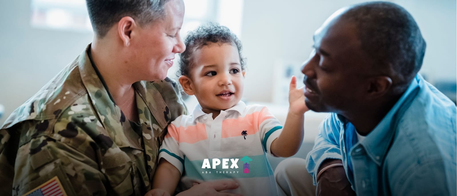 Toddler sitting with a service member in uniform and another adult, reaching toward the adult's face