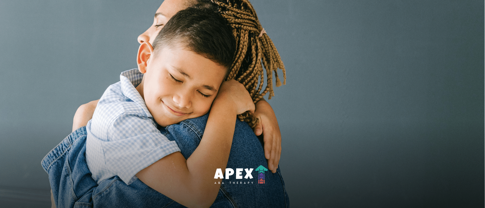 Child hugging an adult with braided hair, eyes closed and smiling, against a gray background.