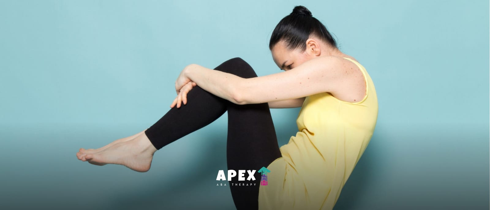 Woman in a yellow top sits curled up with knees drawn to her chest against a teal background, conveying a quiet, introspective mood.