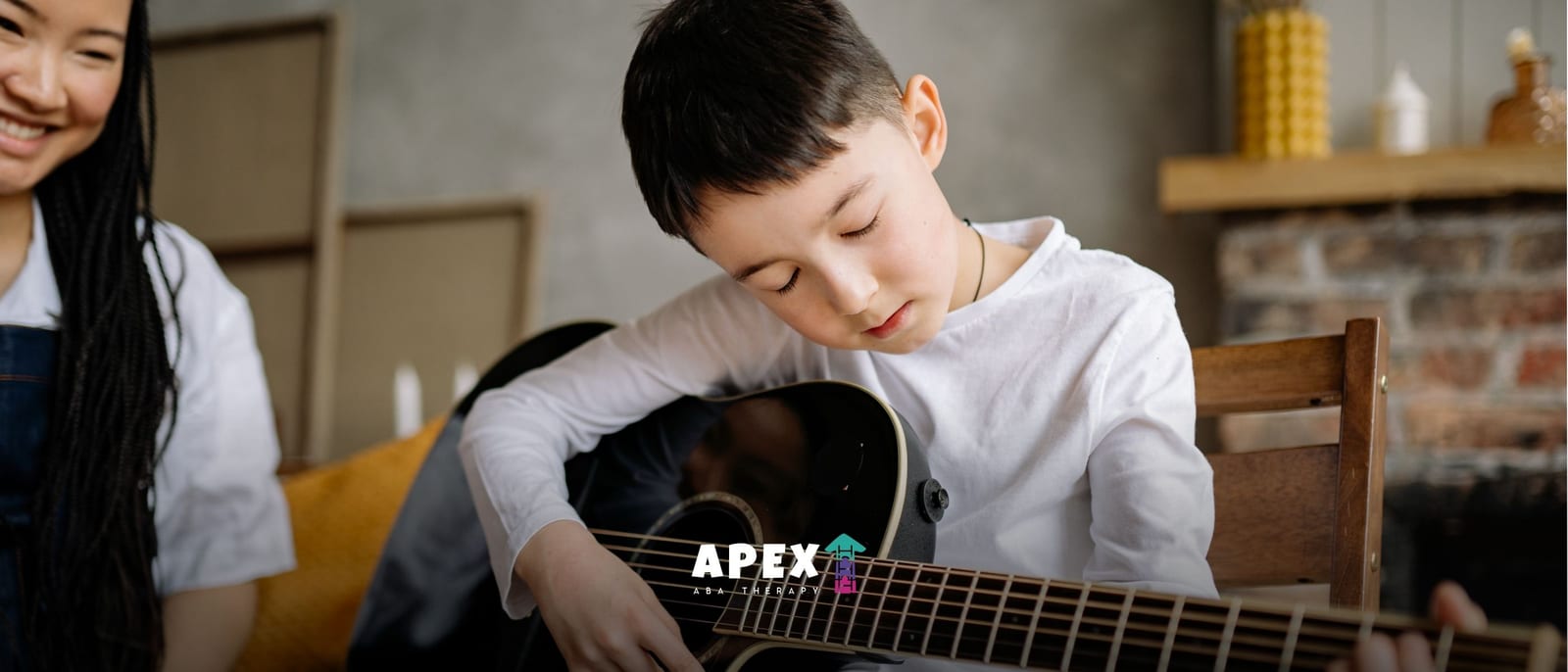 A young boy in a white shirt focuses on playing an acoustic guitar as a smiling woman watches nearby, creating a warm, relaxed music lesson scene.