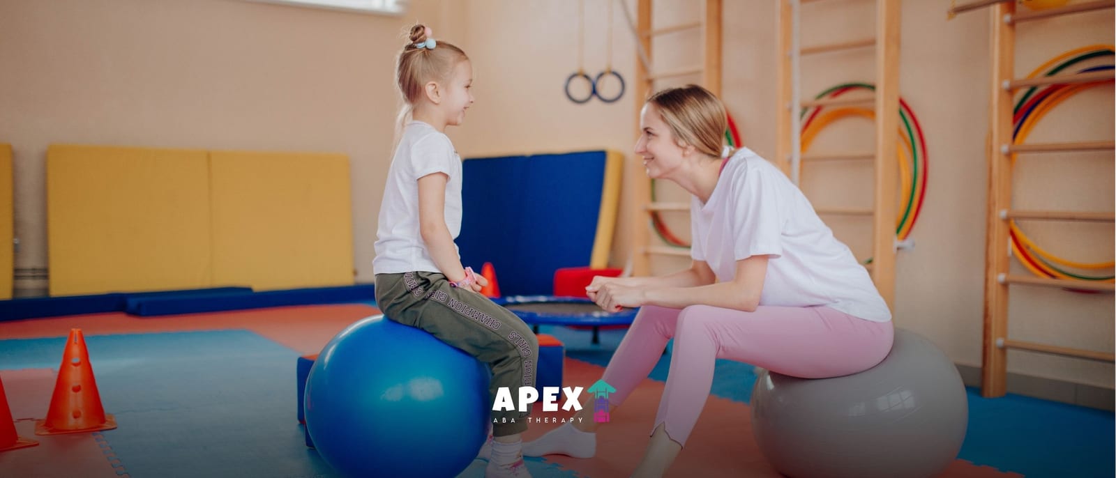 A physiotherapist smiles at a young girl seated on a blue exercise ball in a colorful therapy room, creating a warm, encouraging mood.
