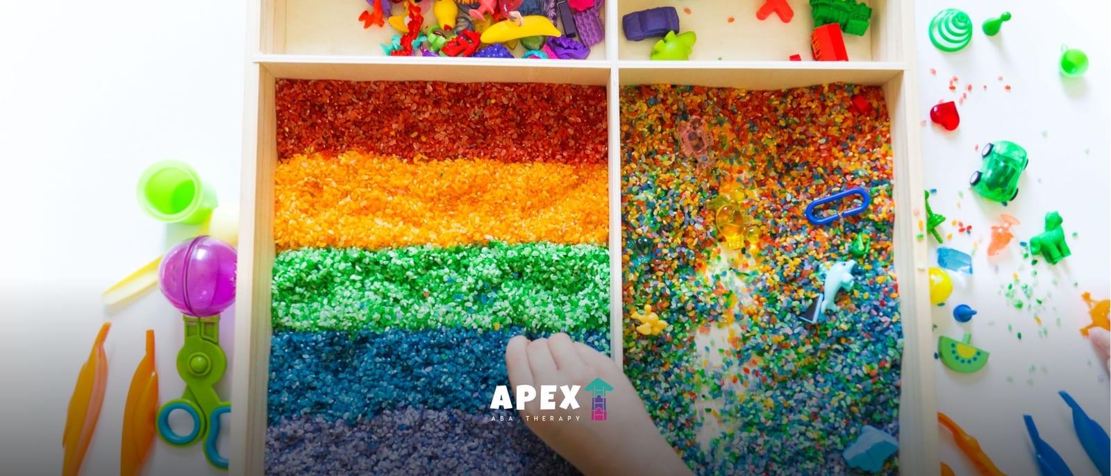A hand reaches into a wooden sensory tray filled with colorful grains and beads, surrounded by scattered toys and craft supplies in a playful setting.