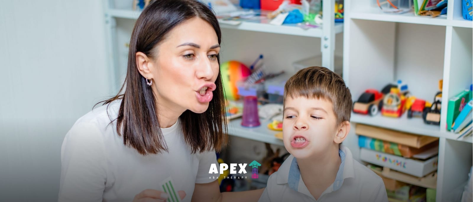 A woman and young boy make playful puckered-lip faces in a bright toy-filled room, with shelves of toys behind them and an APEX logo below.
