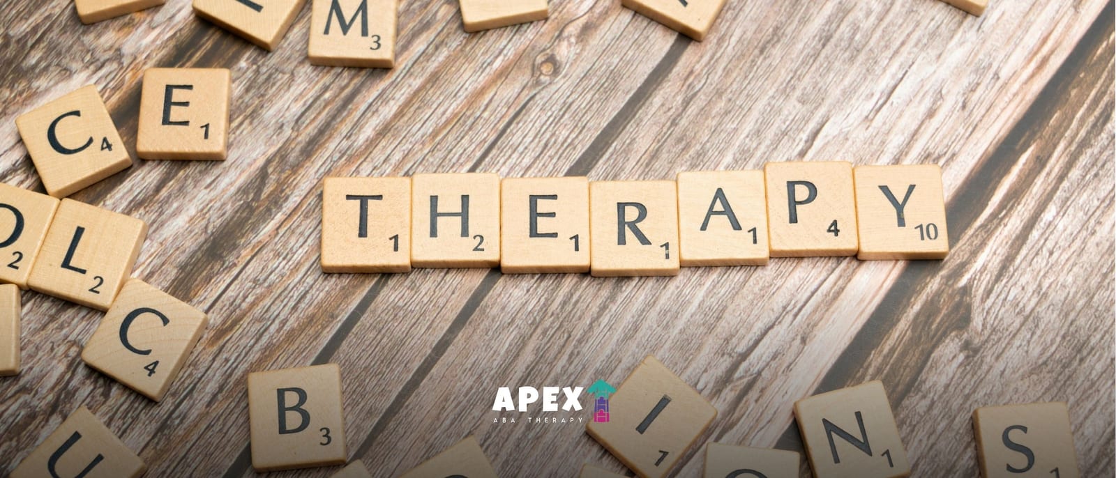 Wooden letter tiles spell “THERAPY” across a weathered tabletop, with scattered Scrabble pieces and an APEX logo below, evoking reflection.