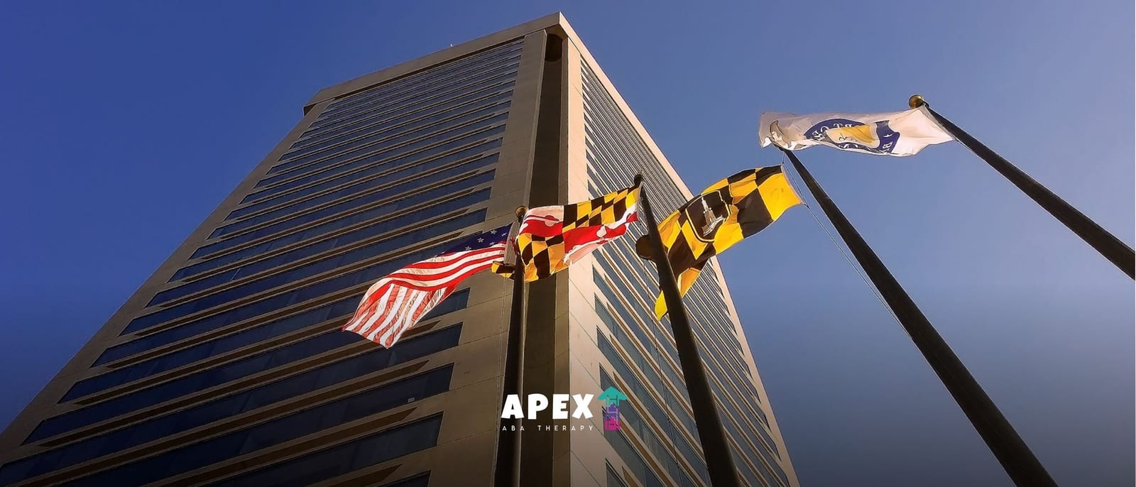 American, Maryland, and Baltimore flags flying in front of a tall office building against a clear blue sky