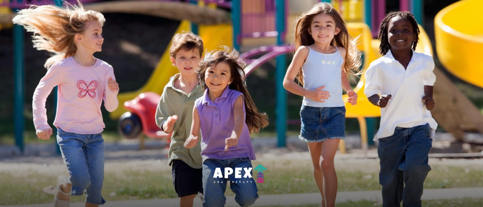 Group of smiling children running together on a playground with colorful play equipment in the background.